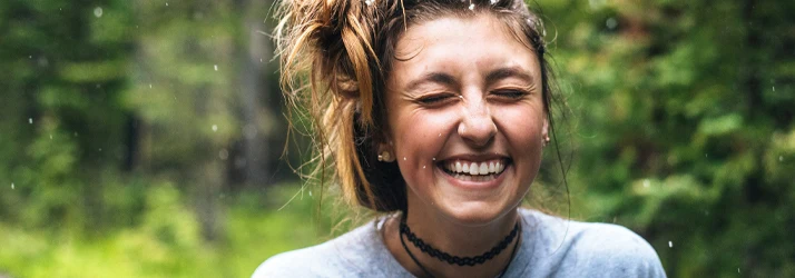 Periodontist Portland ME Young Woman with Hair in a Messy Top Bun is Outside Smiling With Her Eyes Closed Enjoying the Weather Crowns Focused Young Woman with Hair in a Messy Top Bun is Outside Smiling With Her Eyes Closed Enjoying the Weather